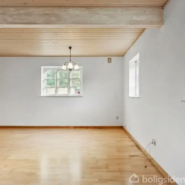 Et tomt rum med trægulv og hvidmalede vægge. En lysekrone hænger ned fra loftet. Værelset har flere vinduer og en trædør på venstre side.