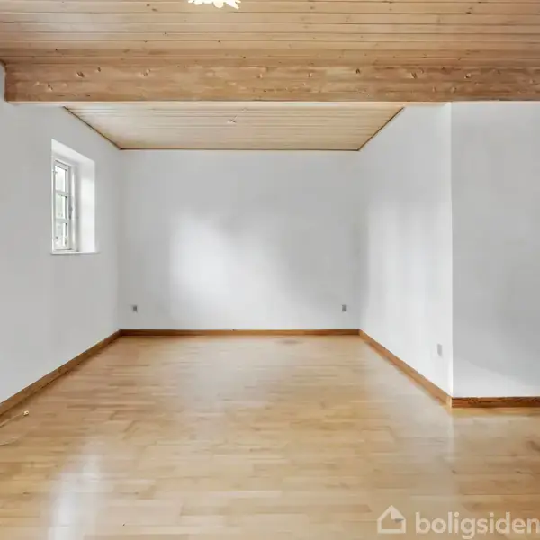 Stue med trægulv strækker sig ud, mens en pejs står stille i hjørnet. Lyse vægge og et loft med synlige bjælker skaber en åben og minimalistisk atmosfære.