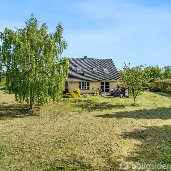Hus står stille i en stor græsplæne med enkelte træer omkring, herunder en hængende pil. Himlen er blå, og der er buske langs kanterne af plænen.