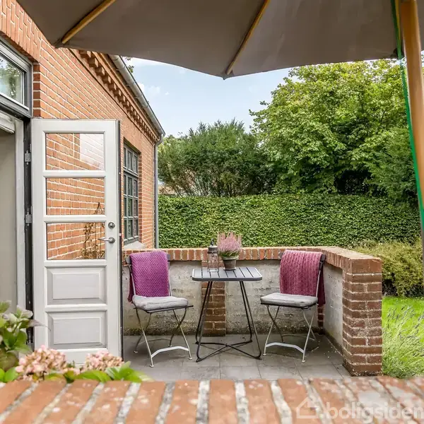 Terrasse med to stole og et lille bord, flankeret af murstensvægge. En åben dør fører ind i huset. Parasol skaber skygge, omgivet af grønne buske og græs.