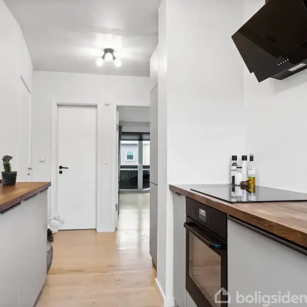 Køkken med træbordplader, integreret ovn og sort emhætte. En lang gang fører til en dør. Der står krydderier på bordet og en kaktus i hjørnet.