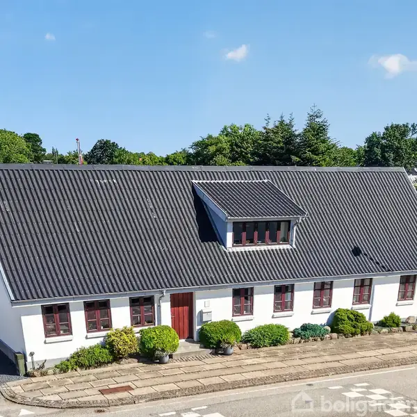 Et hvidt hus med sort tag har flere røde vinduer og en dør langs siden. Det står i en urban kontekst med grønne buske foran og en blå himmel bagved.