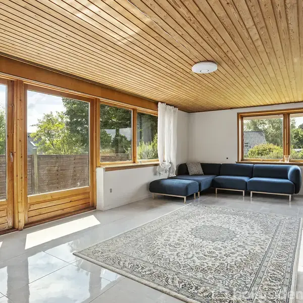 Stue med blå sofa i hjørnet, store vinduer og døre mod haven, trægulv, tæppe på flisegulv, hvidt loft og gardiner.