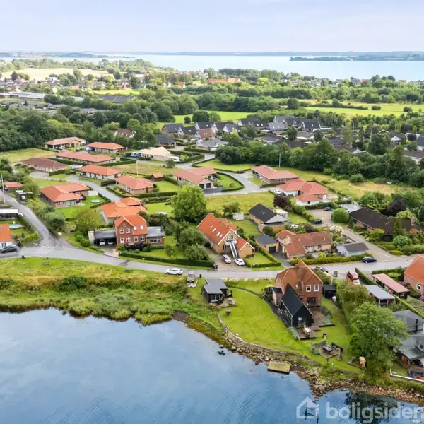 En klynge af huse med røde tage ligger tæt på en sø og grønne områder. I baggrunden ses flere huse og åbent landskab med udsigt til vand.