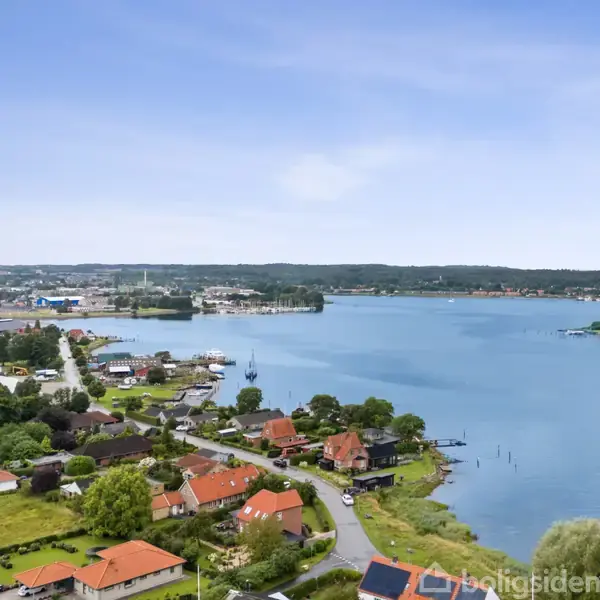 En by med rødt tagede huse ligger langs en buet vej ved en bred, stille flod. Skov og industriområde ses i baggrunden under en klar blå himmel.