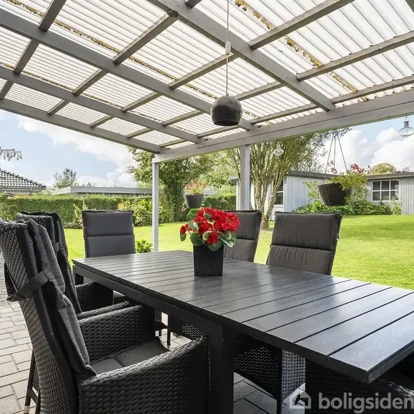 Terrassemøbler står på en overdækket terrasse, med et sort træbord omgivet af kurvestole. Potteplante med røde blomster er på bordet. Græsplæne og haveskur ses i baggrunden.