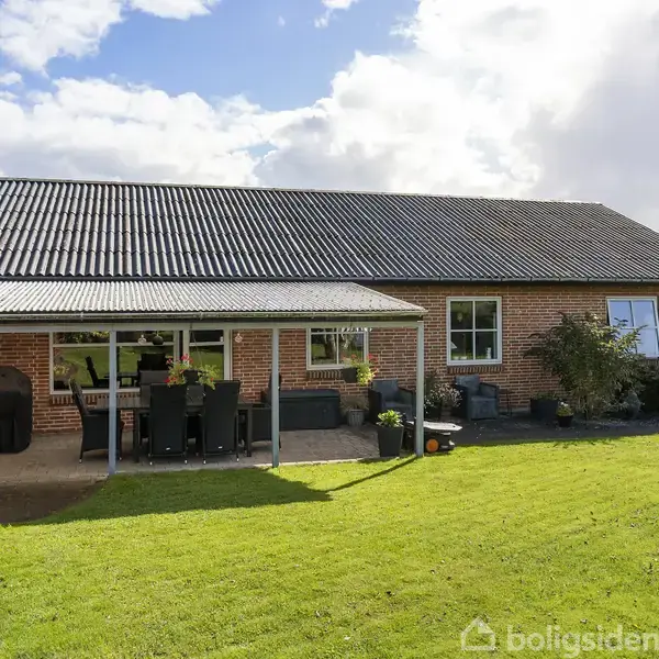 Rødt murstenshus med sort tag, dækket terrasse med havemøbler. Grønt græsplæne omgiver huset, i en rolig forstadsmæssig have. Solskin og skyer i baggrunden.