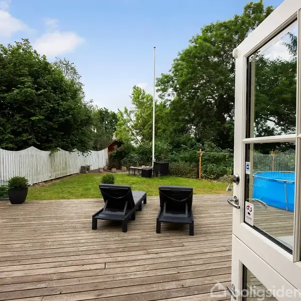 En terrasse med to sorte liggestole vender ud mod en have med grønt græs, blå pool, hvidt stakit og mange træer i baggrunden.