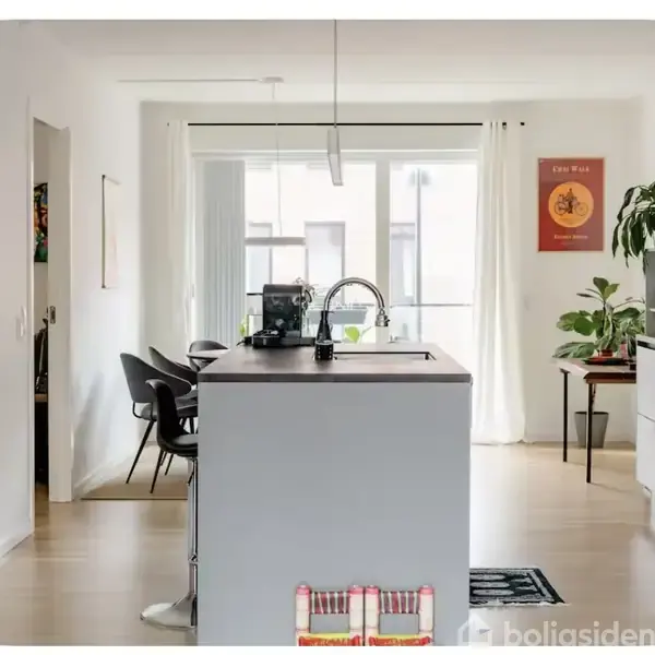 Et moderne køkken har en ø med vask, omgivet af hvide og grå nuancer. En kaffemaskine står på øen. Til højre er der grønne planter og en ovn. Plakaten på væggen har teksten: "The War of Art."