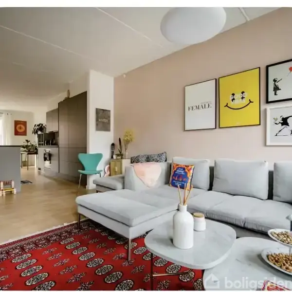 Grå hjørnesofa med puder i moderne stue, dekoreret med kunstplakater på lyserød væg. Rundt sofabord med vaser og snacks. Åbent køkken i baggrunden. Tekst: "FEMALE".