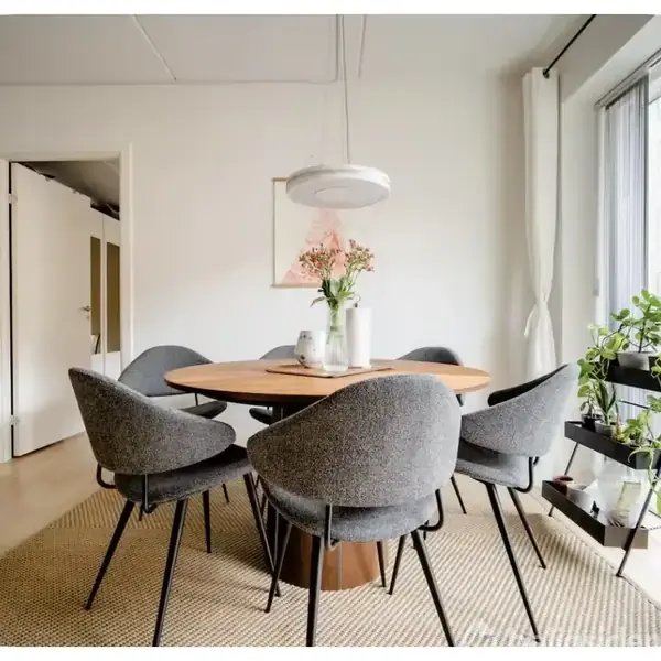Rundt træbord omgivet af seks grå stole, dekoreret med en vase blomster. Ophængt lampe over bordet. Lys indretning med lysindfald fra vindue og grønne planter i hjørnet.