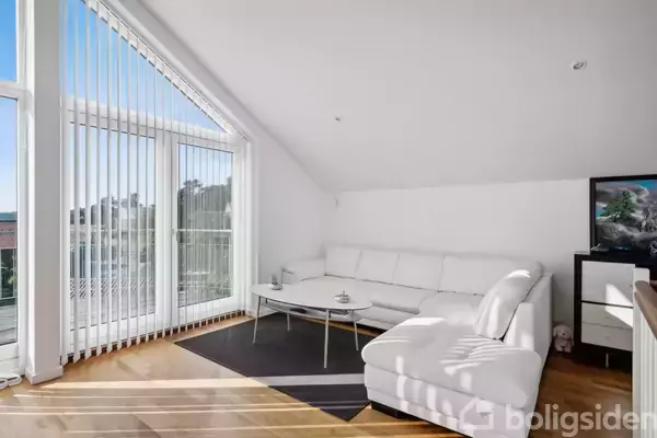 En hvid hjørnesofa står stille i en lys stue med skråvægge. Store vinduer med persienner tillader lysindfald. Et sofabord står foran, og et maleri hænger på væggen.