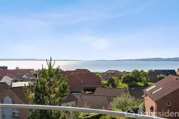 Huse med røde tage står stille i en forstad med udsigt over en rolig sø i baggrunden under en klar, blå himmel.