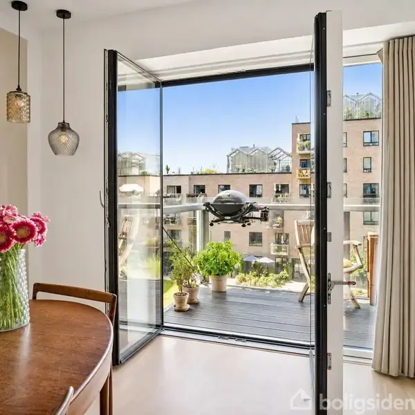 Blomster i vase står på rundt bord i stue. En altandør er åben, viser balkon med planter og udsigt over moderne lejlighedskompleks. Sollys skinner ind, skaber lys og rummelighed.