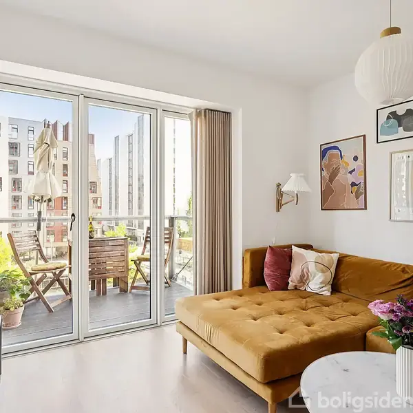 Brun sovesofa står i lys stue, dekoreret med vægkunst. Glasskydedøre fører ud til balkon, hvor der er træbord og stole. Bygninger ses i baggrunden udenfor.