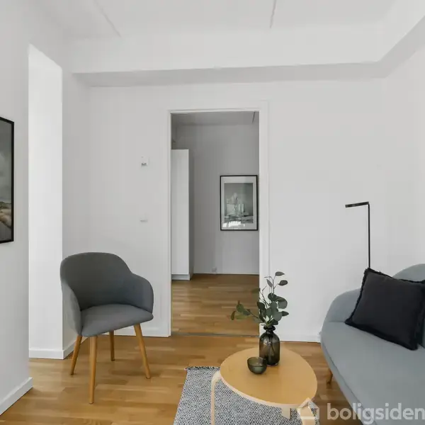En stue med en grå sofa og en grå stol, et lille rundt sofabord med en vase, to fotografier på væggen og parketgulv. I baggrunden ses en åbning til et andet rum.