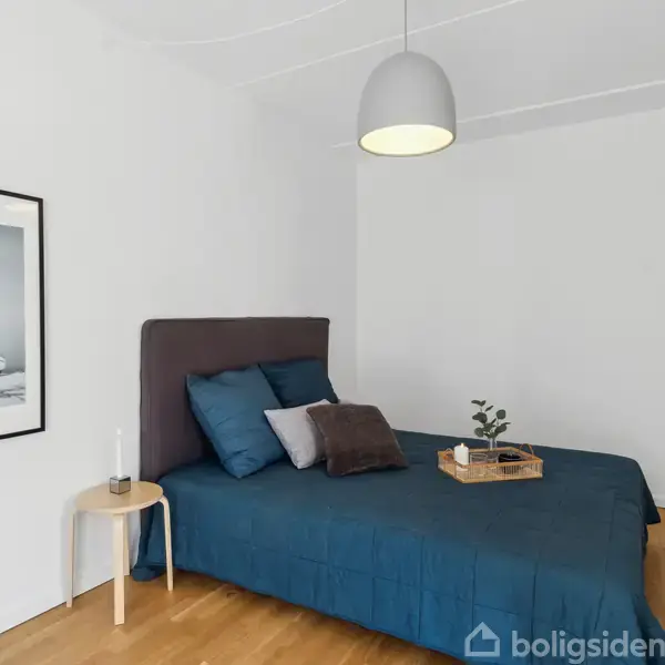 En seng med blåt sengetøj står i et lyst værelse med trægulv. En lille rund natbord holder en lysestage. En plakat af både hænger på væggen.