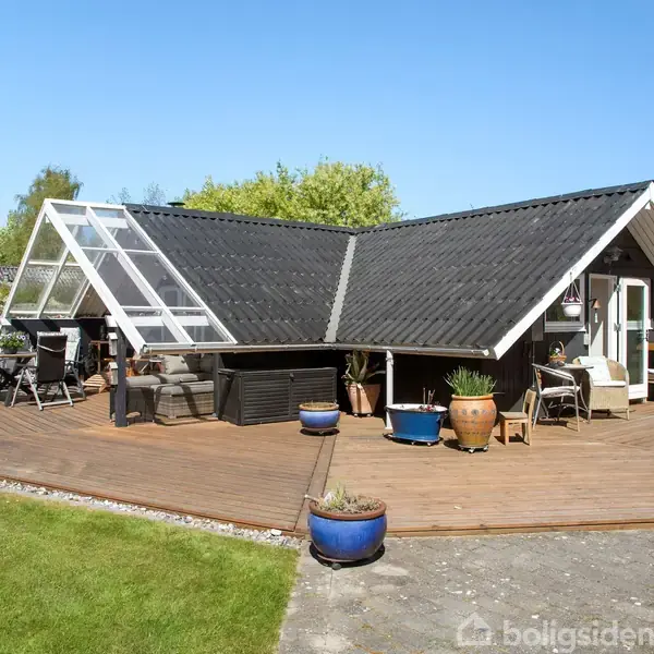 Et sort-hvidt sommerhus med en V-formet tagkonstruktion står på en stor træterrasse. Der omkranses af blomsterkrukker og grønne planter, mens et skifergulv leder ind til græsplænen.