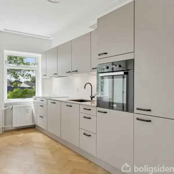 Et moderne køkken med grå skabe, en indbygget ovn og mørk vandhane. Det hvide rum har stort vindue med udsigt til grønne træer. Brunt parketgulv fuldender indretningen.