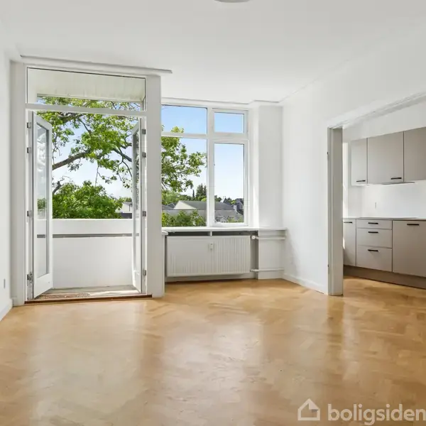 Et lyst rum med parketgulv har en åben dør til en balkon, udsigt til grønne træer. Til højre ses et køkken med grå skabe. Ingen tekst er synlig.