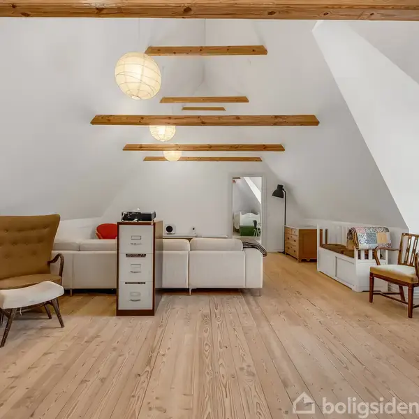 Loftstue med træbjælker i loftet, hvid sofa og lænestol. Baggrunden har en buet vinduesniche med to træstole og en globus. Rummet er lyst med trægulv.