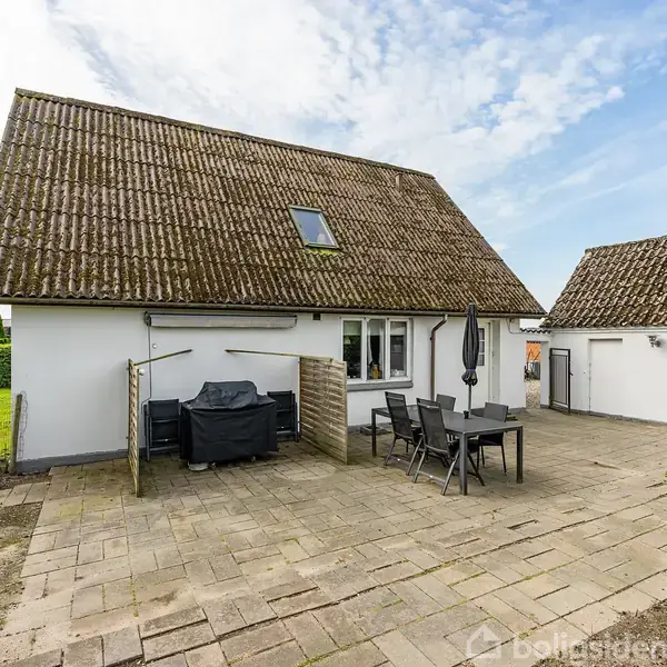 En hvid villa med skrå tag samt kvistvindue. Foran huset står havemøbler og en grill på en flisebelagt terrasse. Haven er omkranset af grønne buske.