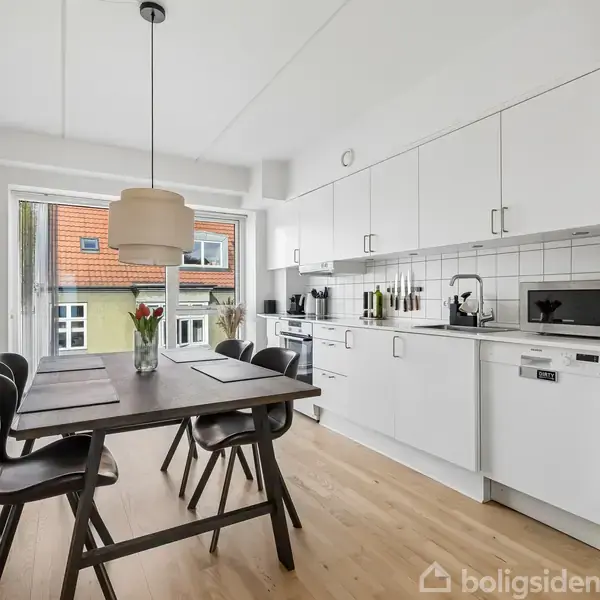 Et moderne køkken med hvidt inventar og trægulv. Et spisebord med fire stole er centralt placeret. En stor lampe hænger over bordet. Store vinduer giver naturligt lys.