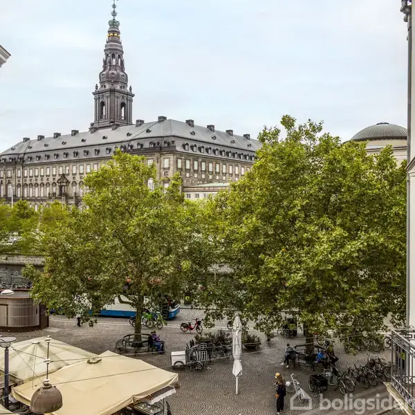 En stor historisk bygning med tårn ses gennem frodige træer. I forgrunden er en åben plads omgivet af bygninger, med caféborde og mennesker der sidder og nyder omgivelserne.