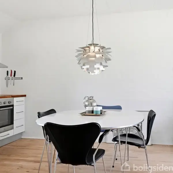 Et ovalt spisebord står i et moderne køkken med fire stole omkring, en hængende lampe over bordet, og en guitar læner sig mod væggen ved siden af en lænestol.