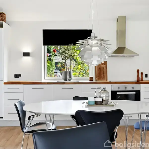 Et rundt spisebord er omgivet af stole i et moderne køkken. Køleskabet og ovnen er indbygget i hvide skabe. Et vindue med udsigt til en have giver lys.