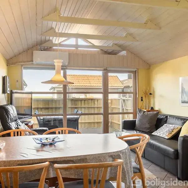 Et spisebord med stole står i centrum, en sofagruppe til højre. En lænestol er placeret ved glasdøren, som fører ud til en terrasse. Lyse vægge og skrå loftsbjælker omkranser rummet.