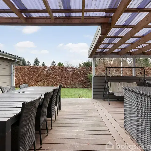 Der er et overdækket træterrasse med et stort spisebord og stole. Terrasseområdet har også en sofasæt og en hængesofa, og er omgivet af en hæk og græsplæne.