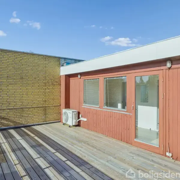 Træterrasse foran et rødt træhus med store vinduer, beliggende ved en gul murstensvæg. En hvid luftkonditioneringsenhed hænger på væggen. Klar blå himmel ses i baggrunden.