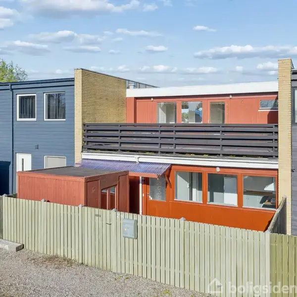 To-etagers rækkehus med en grå og rød facade ses bagved et lavt hegn. Et fladt tag og store vinduer præger bygningen. Skyer og trætoppe ses i baggrunden.