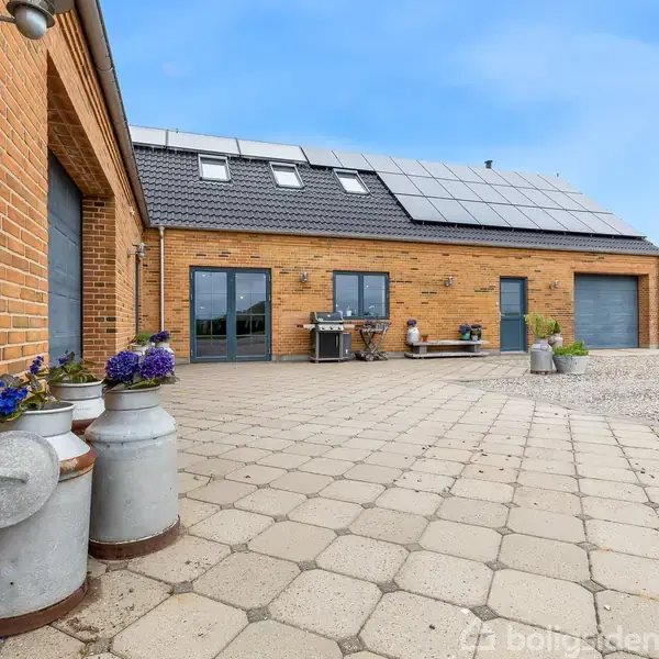 En gårdsplads med fliser og mælkespande i forgrunden. I midten står en grill og et bord. Huset har solpaneler på taget. Omkringliggende natur i baggrunden.