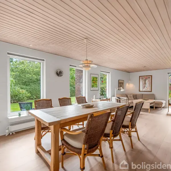 Spisebord omgivet af seks stole står i en lys, moderne stue med store vinduer, der viser grønne omgivelser udenfor. Baggrunden indeholder en sofagruppe og træpanel-loft.