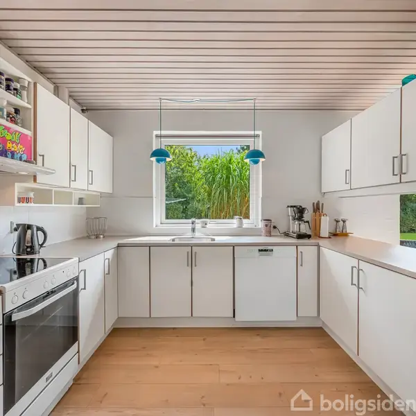 Køkken med hvide skabe og træbordplader. Komfur og elkedel står på venstre bord. Vindue over vasken med udsigt til have. Hylde med krydderidåser øverst.