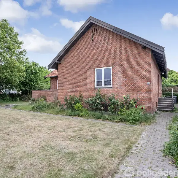 Rødt murstenshus står stille ved en have med græsplæne og en flagstang. Brostensbelagt sti fører op til huset, omkranset af grønne buske og træer under en blå himmel.