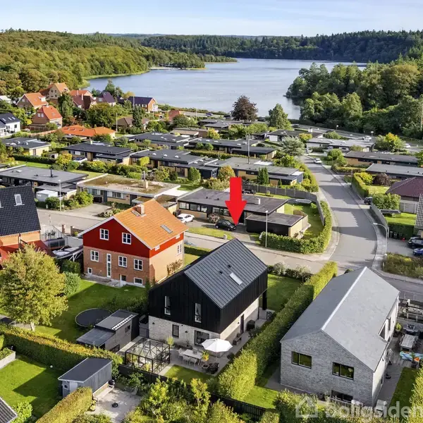 Rødt hus står blandt moderne boliger. En rød pil peger på det fra luften. Omgivet af grønne haver med udsigt til skov og sø i baggrunden.
