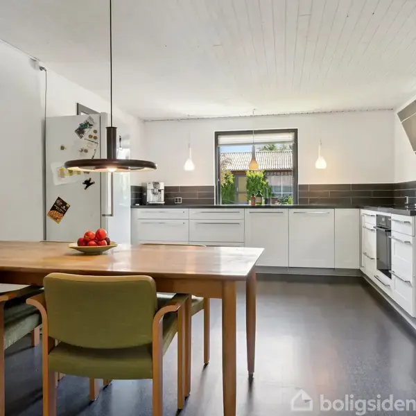 Et moderne køkken med et træbord og to grønne stole i forgrunden. Køkkenet har hvide skabe, sort bordplade, og stålapparater. Vinduer slipper naturligt lys ind. En skål med frugt står på bordet.