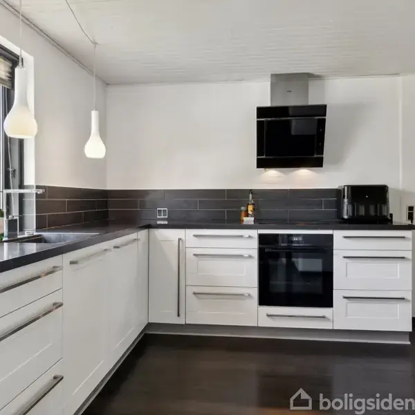 Hvidt køkken med mørke bordplader, indbygget ovn og emhætte. Vinduet til venstre giver udsigt til haven. To hængende lamper og en kaffemaskine står på bordet.