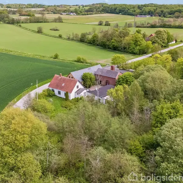 To huse ligger omkranset af frodig grønt landskab og marker. En snoet vej fører gennem området. Omkringliggende skove og åbne områder skaber en fredelig landlig scene.
