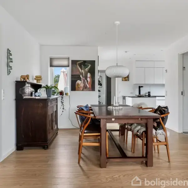 Spisebord med stole omkring står på trægulv i et åbent rum; et køkken med hvide skabe ses i baggrunden. En stor plante og kunstmaleri tilføjer dekoration.