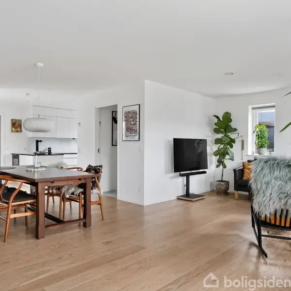 Stue med spisebord og stole, der står på lyse træplanker. I baggrunden ses et åbent køkken med hvidt interiør. Planter og kunstværker præger rummets lyse og moderne indretning.