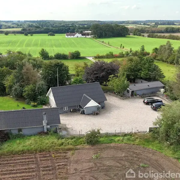 Et parcelhus med sort tag står stille omgivet af grønne marker og træer. Ved siden af er en parkeringsplads med flere biler. Landsbynatur spreder sig i baggrunden.