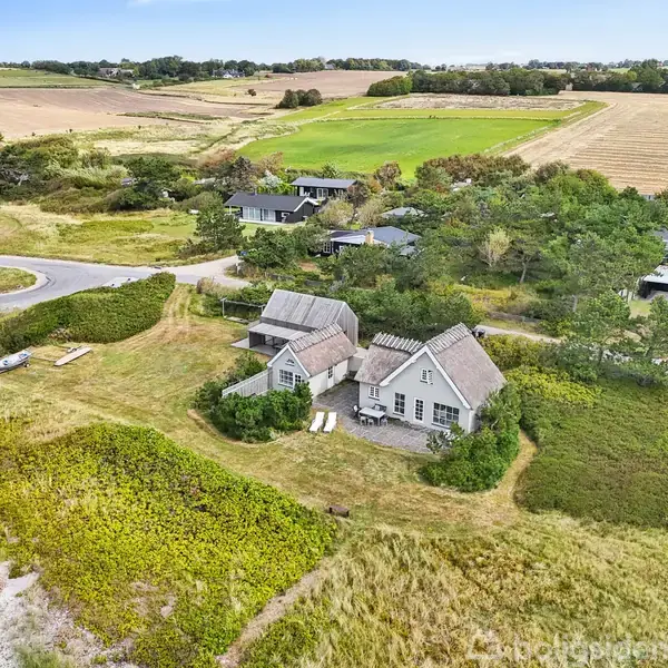 Et hus med to sammenhængende bygninger ligger i en åben grøn mark tæt på en strand. Omkring huset ses flere mindre huse og en vej med parkerede biler.