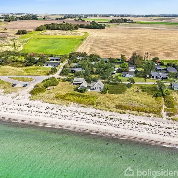 Strandhuse ligger stille langs kysten, omgivet af grønne marker og klare, blå bølger, med spredte boliger og adgangsveje mellem naturen.