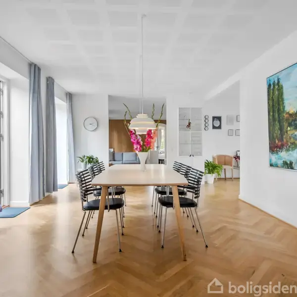 Et lyst spisebord med stole omkring står i en elegant spisestue med trægulv. Der hænger en stor maleri på væggen og vinduer med blå gardiner åbner mod en balkon.