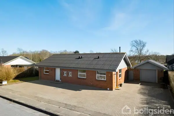 Rødt murstenshus står stille på en flisebelagt indkørsel, omgivet af en garage og hække. Blå himmel og træer ses i baggrunden.