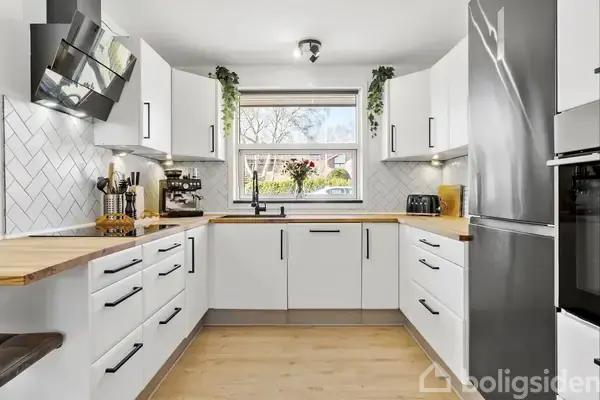 Et moderne køkken med hvide skabe og sorte håndtag er fyldt med apparater. En stor vindue bagved afslører udsigt til træer og buske. Planter pynter rummet.